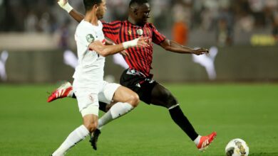 Two soccer players chase the ball on a green field, one in white and one in red-and-black, reaching for a low kick as the ball rolls ahead.