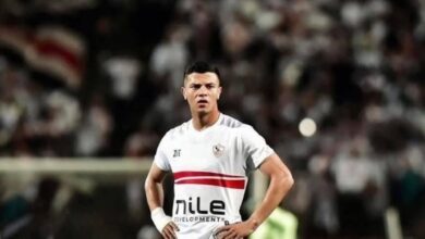 Soccer player in a white jersey with a red stripe, standing with hands on hips on a stadium field, blurred crowd behind him.