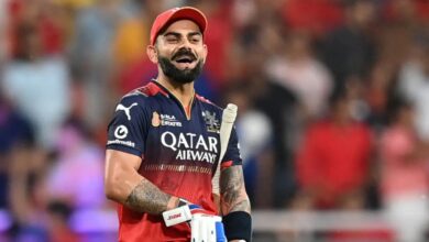 Cricketer in a red and navy jersey holding a bat, smiling on the cricket field with a blurred crowd in the background.