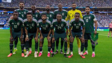 Saudi national football team players pose for a group photo on the pitch before a match, in green jerseys with numbers visible, goalkeeper in yellow.