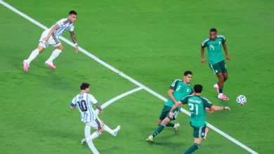 Argentine players in light-blue striped uniforms contest the ball against four green-clad defenders on a green pitch as the ball bounces nearby.