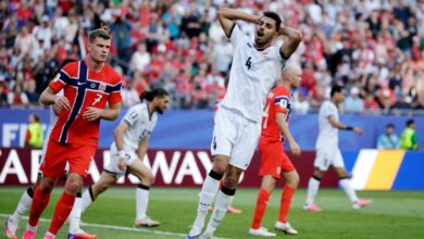 Soccer player in white #4 holds his head with hands, looking surprised as he runs on the field during a match with red-jersey opponents and a crowded stadium behind him