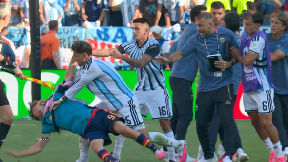 Soccer match scuffle: players from two teams tug at a downed opponent on the field while teammates and officials intervene, crowd watching in the background.