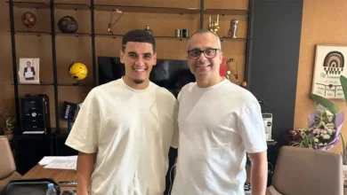 Two men in white t-shirts smiling and posing together in a cozy office/photo studio setting with shelves and awards in the background.