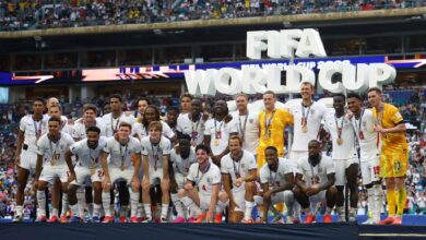 England football team posing on a podium with medals after winning World Cup, large 3D WORLD CUP letters in the background and a cheering stadium crowd.