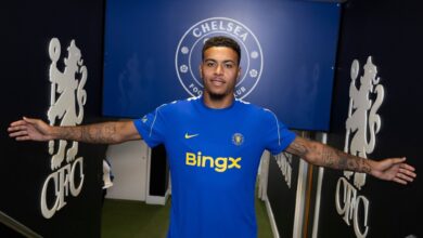 Chelsea FC player in a blue jersey stands with arms outstretched in a tunnel, the Chelsea crest visible behind him on a blue wall.